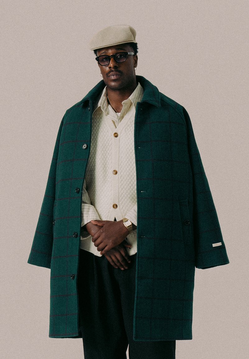 Grønn rutete frakk med en strukturert design, med krage og knapper, lagdelt over en quilted kremvest og sorte bukser.