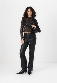 Schwarzes, transparentes Langarmoberteil mit einem Muster von Halbmond; eng anliegende schwarze Lederhosen; spitz zulaufende schwarze Stiefel; kleine schwarze Handtasche.