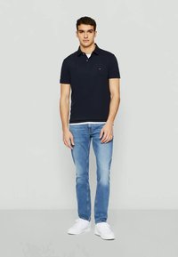 Camisa polo azul-marinho com mangas curtas, tecido leve e um pequeno logo no peito. Combinada com jeans azul-claro e ténis brancos.