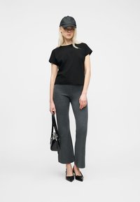 Femme portant une casquette noire, un t-shirt noir, un pantalon évasé gris, tenant un sac à main noir et des chaussures pointues noires sur un fond blanc.