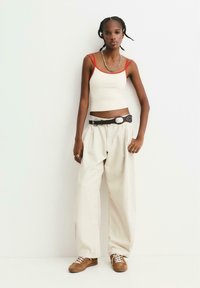 Jonge vrouw met gevlochten haar, gekleed in een witte cropped top met rode bandjes, losse beige broek, zwarte riem en bruine sneakers, staand.