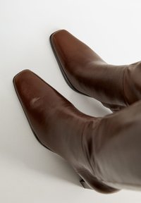 Bottines en cuir marron avec un bout carré, texture lisse, tige fine à la cheville et petit talon carré, sur un fond neutre.