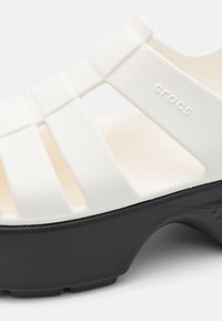 Sandale Crocs blanche avec semelle noire et trois larges brides sur le dessus, montrant un matériau texturé et le nom de la marque embossé.
