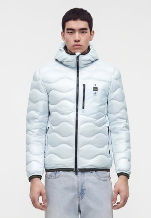 Jeune homme portant une veste matelassée bleu clair avec capuche, fermeture éclair noire et jean délavé clair sur fond blanc uni.