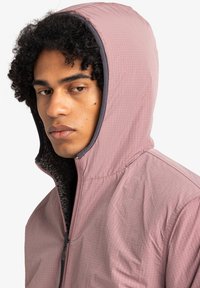 Veste à capuche rose clair avec une fermeture éclair à l'avant, tissu extérieur texturé et intérieur doublé en polaire. Finitions grises le long de la capuche.