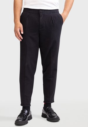 Mann trägt schwarze plissierte Hose, schwarze Socken, schwarze Lederloafer und weißes Hemd, steht vor einem schlichten Hintergrund.