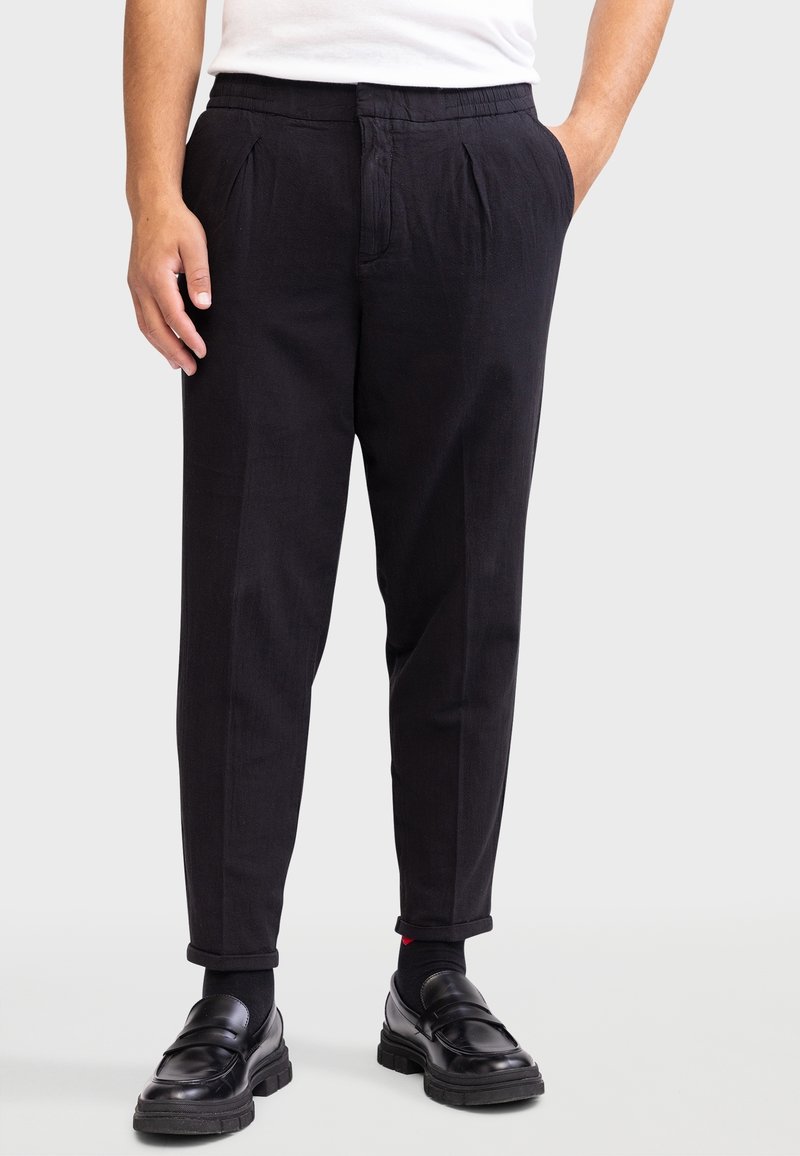 Homme portant un pantalon plissé noir, des chaussettes noires, des mocassins en cuir noir et une chemise blanche, debout devant un fond uni.