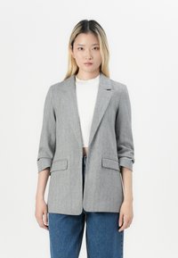Grijze blazer met houndstoothpatroon, met een enkelvoudige sluiting, twee voorzakken en opgerolde mouwen. Gecombineerd met high-waisted jeans.