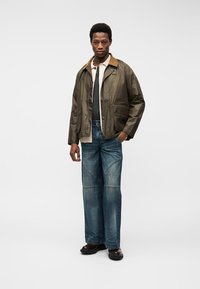 Homme debout portant une veste marron avec un col beige clair, une chemise beige clair, un sous-pull gris foncé, un jean bleu et des chaussures marron foncé.