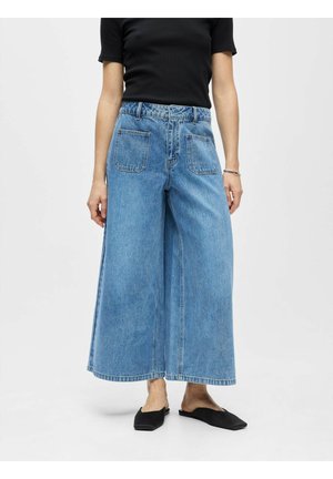 Vid bensnitt, croppede blå jeans med forlommer, brukt med svart topp med korte ermer og svarte flate slip-on-sko.