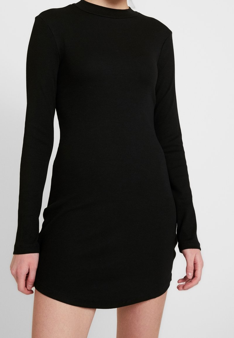 Femme portant une robe mini noire moulante à manches longues avec un col rond haut sur un fond clair uni.