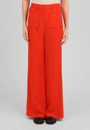 Pantalon rouge taille haute à jambes larges avec grandes poches plaquées à l'avant, porté avec des chaussures tissées noires et un haut beige sans manches.