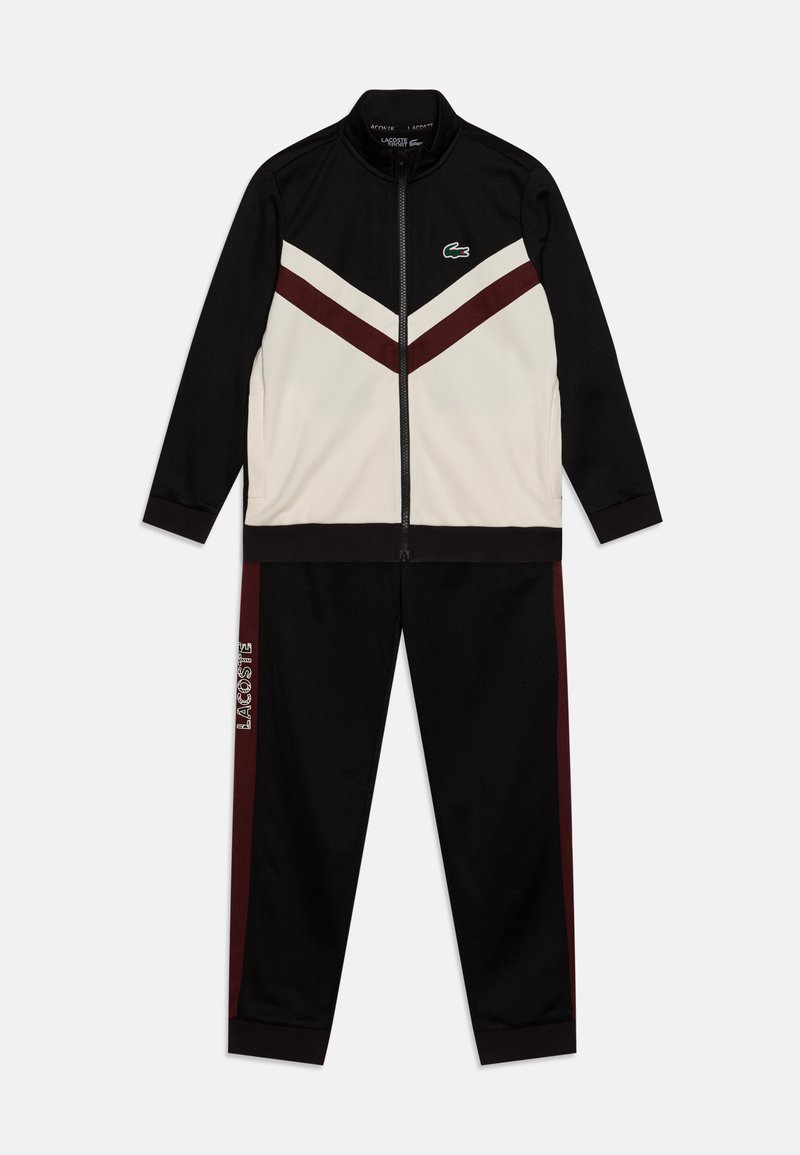 Ensemble de survêtement noir et crème avec une veste zippée au design en chevrons, et un pantalon assorti avec des rayures bordeaux et un logo. Texture de tissu lisse.