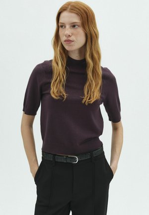 Joven con pelo largo y rojo que lleva una camiseta de manga corta color púrpura oscuro y pantalones negros con las manos en los bolsillos, mirando hacia un lado.