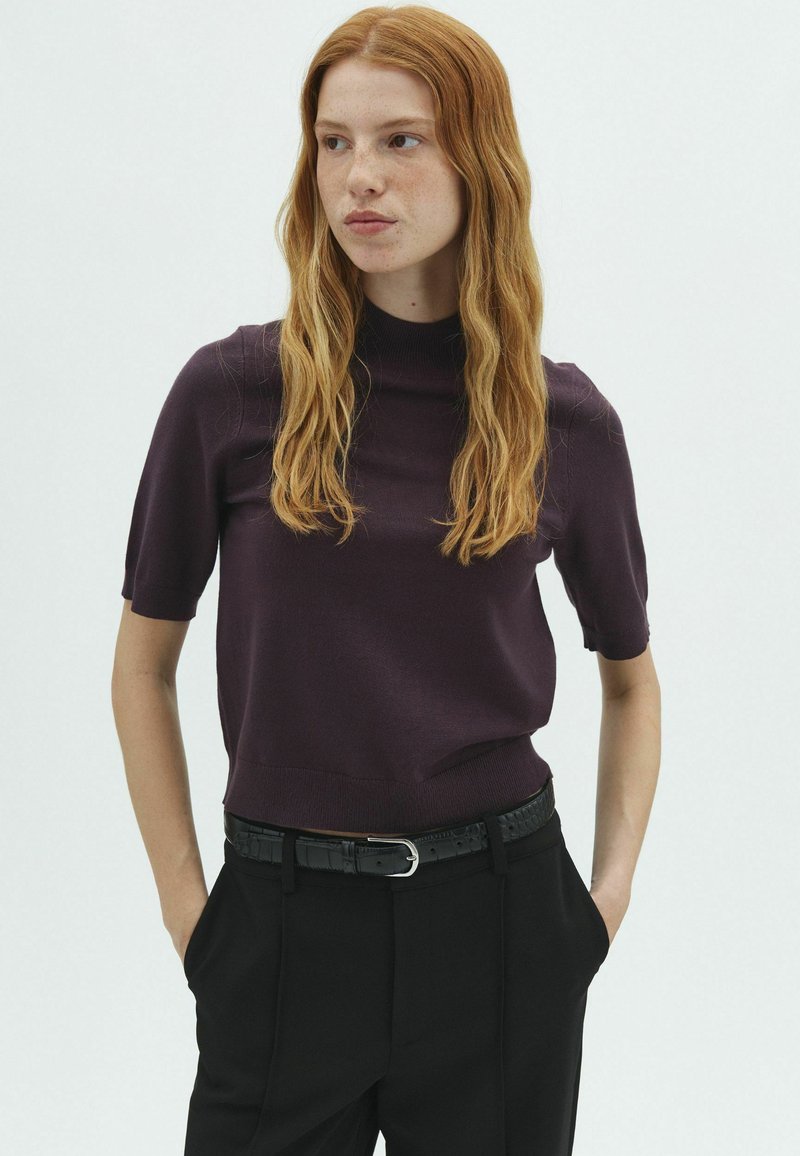 Jeune femme aux longs cheveux roux portant un haut violet foncé à manches courtes et un pantalon noir, les mains dans les poches, regardant sur le côté.