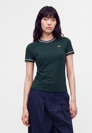 Femme aux longs cheveux noirs portant un t-shirt vert foncé à manches courtes avec une bordure claire et un pantalon ample bleu marine, debout devant un fond uni.