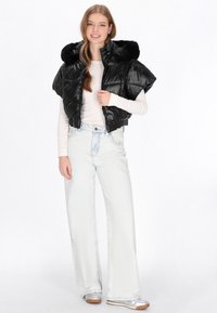 Czarna pikowana kamizelka z kapturkiem z sztucznego futra, krótki krój, błyszcząca tekstura i szerokie ramiona. Połączona z jasnoniebieskimi dżinsami z szerokimi nogawkami i srebrnymi butami.