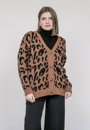 Femme portant un cardigan boutonné imprimé léopard marron et noir sur un col roulé noir, assorti à un pantalon noir, debout devant un fond uni.