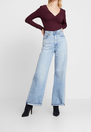 Kobieta w jasnoniebieskich dżinsach z wysokim stanem i szerokimi nogawkami, ciemnofioletowej bluzce z długim rękawem, marszczonym dekoltem w serek oraz czarnych wysokich botkach na obcasie z ostrym noskiem.