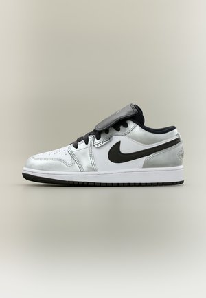 Baskets basses blanches et argentées avec le swoosh Nike noir, lacets noirs, col rembourré et semelle noire sur un fond neutre.