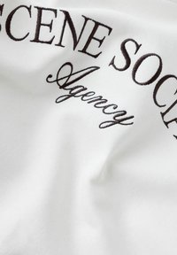 Tela blanca con texto bordado en marrón que dice "SCENE SOCIETY Agency", mostrando una textura suave y un estilo de letra distintivo.