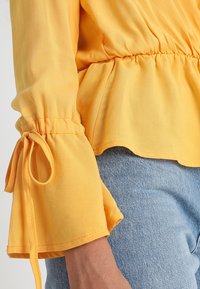 Blusa gialla leggera con vita arricciata, maniche a sbuffo con lacci e texture liscia abbinata a jeans in denim blu chiaro.