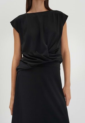 Kvinde iført en sort ærmeløs draperet top, stoppet ind i et sort nederdel med høj talje, stående mod en ensfarvet lys baggrund.