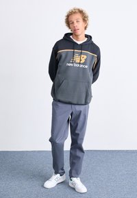 Sudadera con capucha gris con detalles en naranja y bolsillo frontal, combinada con pantalones grises y zapatillas blancas con acentos verdes.