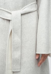 Manteau gris clair en mélange de laine avec ceinture, présentant une texture douce et des manches larges. Fente notable à l'avant pour un style accentué.
