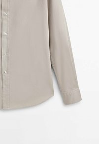 Jasnobeżowa koszula z długim rękawem wykonana z gładkiej tkaniny, z zapięciem na guziki z przodu i zaokrąglonym dołem. Na rękawie widoczny jest szczegół mankietu.