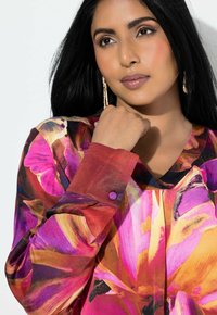 Kvinde med langt sort hår iført guldbøjler og en livlig blomstret bluse i pink, orange og lilla nuancer, poserer med hånden nær hagen.