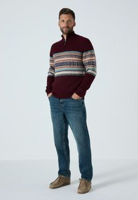 Bordeaux gebreide trui met een halve rits, voorzien van een kleurrijk geometrisch patroon op de borst, gecombineerd met blauwe jeans en bruine schoenen.