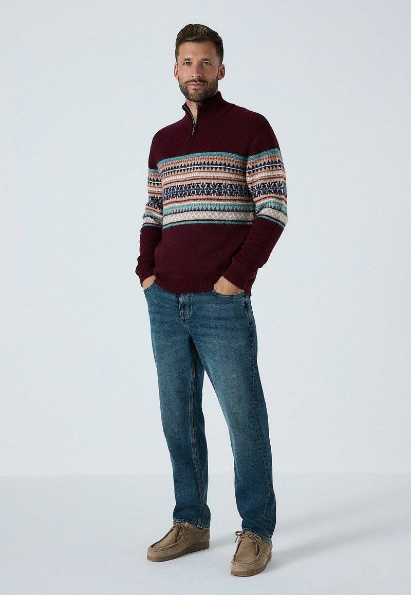 Burgunderfarbener Strickpullover mit halber Reißverschluss, der ein mehrfarbiges geometrisches Muster über die Brust hat, kombiniert mit blauen Jeans und braunen Schuhen.