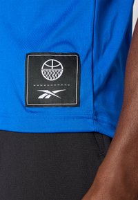 Zila sporta krekls ar melnu kvadrātveida uzšuvi, uz kuras attēlots basketbola grafiks un logo, ar gludu tekstūru un īsām piedurknēm.