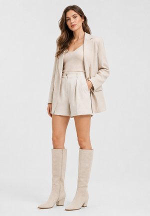Frau mit langen braunen Haaren, die einen beigen Blazer, passend dazu hoch taillierte Shorts, ein Stricktop und kniehohe Stiefel trägt, steht mit einer Hand in der Tasche.