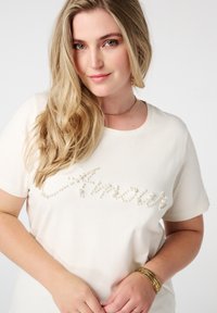 T-shirt en coton couleur crème avec le mot "Amour" orné de petites perles ressemblant à des perles. Manches courtes et encolure ronde.