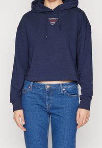 Felpa corta blu navy con cappuccio regolabile e logo "TOMMY JEANS 1985" in rosso sul petto, abbinata a jeans in denim blu.
