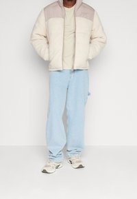 Krämsfärgad och beige fluffig jacka med dragkedja, med en slät övre del och rund krage. Bärs över en ljus skjorta och blå jeans.