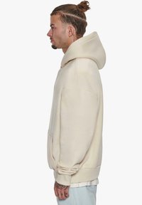 Beige oversized hoodie made of soft fabric, featuring a kangaroo pocket, ribbed cuffs, and a drawstring hood, shown from the side.