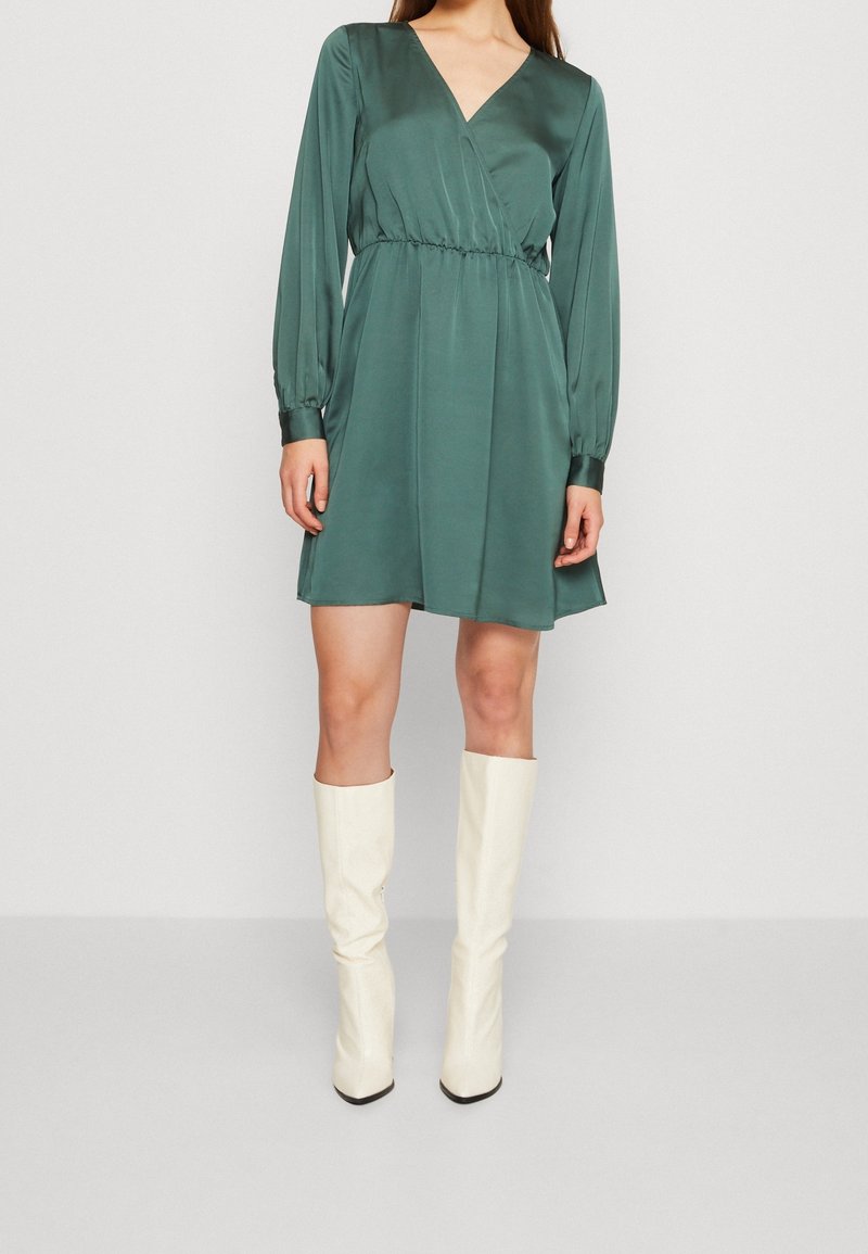 Femme portant une robe vert foncé à manches longues longueur genou avec un décolleté en V et des bottes blanches montantes, debout devant un fond uni.