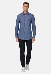 Herenoverhemd met lange mouwen van donkerblauwe stof met een subtiele textuur, knoopsluiting aan de kraag en witte knopen, gecombineerd met een zwarte broek en witte sneakers.