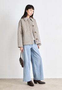 Giacca trapuntata a scacchi in beige e marrone, con grandi bottoni e due tasche frontali, abbinata a jeans a gamba larga azzurri e stivali marroni.