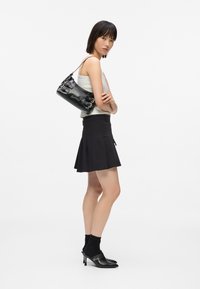 Γυναίκα που φοράει λευκό στενό μπλουζάκι, μαύρη πλισέ φούστα, μαύρες τακούνι μούλες με κάλτσες και κρατάει μια μαύρη δερμάτινη τσάντα ώμου με κούμπωμα.