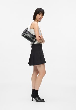 Kvinde iført en hvid tanktop, sort plisseret nederdel, sorte hælede mules med sokker og bærende en sort læder skuldertaske med spænder.