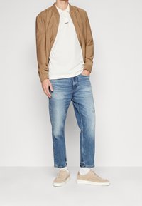 Chaqueta bomber marrón sobre una camiseta polo blanca, combinada con jeans azul claro y zapatillas beige. Texturas suaves y ajuste casual.
