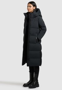 Lange zwarte pufferjas met een capuchon, voorzien van een knoop- en ritssluiting, zijzakken en een geborduurd ontwerp. Gecombineerd met zwarte enkellaarzen.