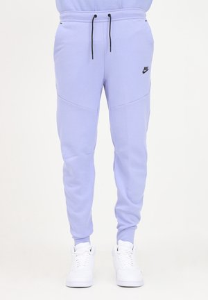 Lyse lilla Nike joggers med sorte snore, sidelommer og tilspidsede ankler båret med hvide sneakers på en ensfarvet baggrund.