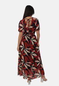 Frau in einem schwarzen Maxikleid mit roten und beigen Blumenmustern, kurzen Puffärmeln, einem gebundenen offenen Rücken und Sandalen mit transparenten Absätzen.