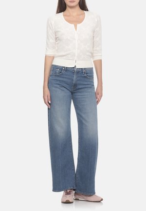 Donna che indossa un cardigan bianco abbottonato, jeans blu a gamba larga e sneakers rosa, in piedi davanti a uno sfondo semplice.