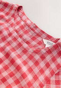 Pehmeästä kankaasta valmistettu punainen ja vaaleanpunainen ruudullinen paita, jossa on pyöreä pääntie ja hillityt ompeludetaljit, näkyvällä merkkilapulla.